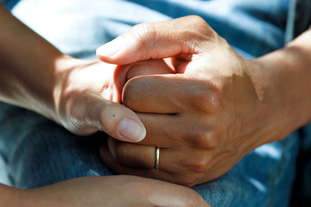 Zwei Hände, die sich in einem zärtlichen Moment umarmen. Eine Hand ist mit einem schlichten Ring geschmückt, der die Schönheit der Verbundenheit einfängt. Der unscharfe Hintergrund erinnert subtil an einen friedlichen Tag in Aachen.