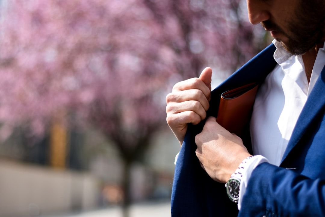 Eine Person im blauen Anzug steckt einen braunen Gegenstand in die Jackentasche, im unscharfen Hintergrund sind rosa blühende Aachener Bäume zu sehen.