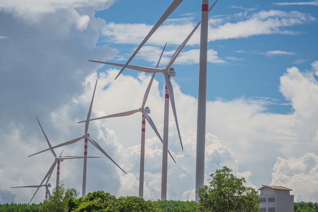 Vor einer Kulisse aus Wolken und blauem Himmel stehen mehrere Windräder, umgeben von Grün in der Nähe von Aachen. In der unteren rechten Ecke ist ein kleines Gebäude zu sehen.