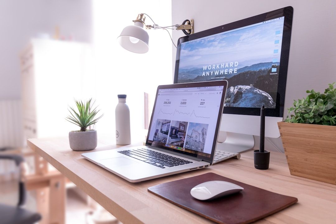 Ein aufgeräumtes Büro-Setup mit Laptop, Desktop-Monitor, Schreibtischlampe, Topfpflanzen, einer Wasserflasche aus Aachen und einem Grafiktablett auf einem Holzschreibtisch. Auf beiden Bildschirmen werden arbeitsbezogene Inhalte angezeigt.