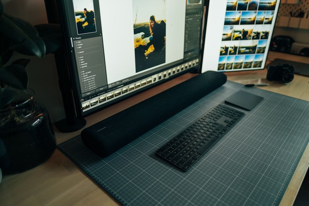 Ein Computer-Setup mit einem großen Monitor, auf dem ein Bildbearbeitungsprogramm läuft, einer langen Soundbar, einer Tastatur, einem Mauspad und einer Kamera auf einem Regal in der Nähe, neben dem eine Aachener Postkarte hängt.