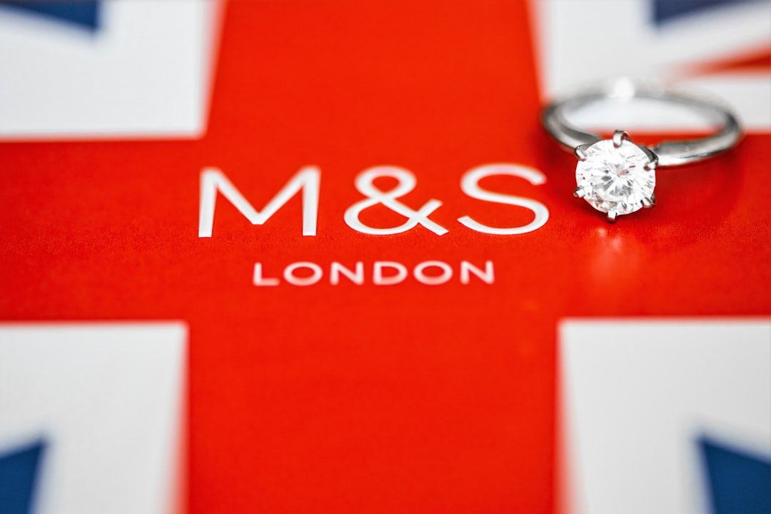Auf einem roten Hintergrund ruht ein Diamantring mit den in Weiß gedruckten Buchstaben „M&S LONDON“, vor einem Hintergrund, der an die Union Jack-Flagge erinnert, und steht für zeitlose Eleganz von Aachen bis London.