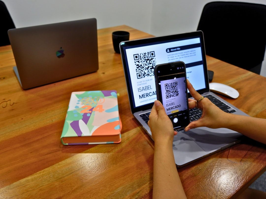 Eine Person scannt mit einem Smartphone einen QR-Code von einem geöffneten Laptop-Bildschirm auf einem Holztisch in Aachen. Auf dem Tisch liegen außerdem ein zugeklappter Laptop und ein bunter Planer.
