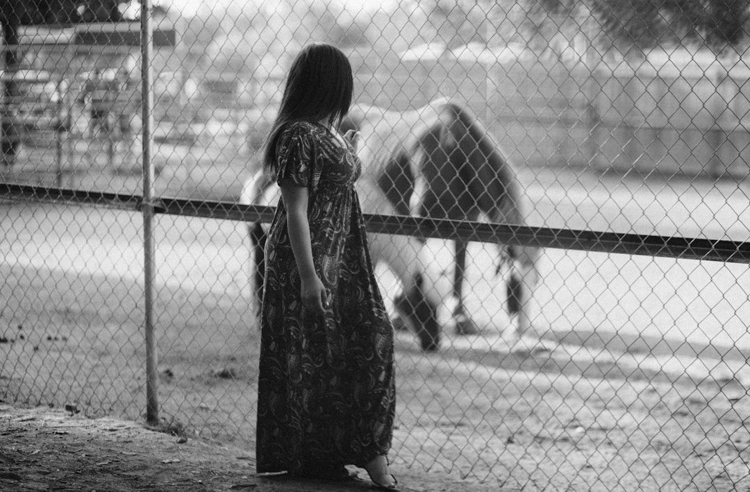 Eine Person in einem langen Kleid steht an einem Maschendrahtzaun und blickt auf ein Pferd hinter dem Zaun. Die Schwarz-Weiß-Szene ruft ein zeitloses Gefühl hervor, das an alte Fotografien aus Aachen erinnert.