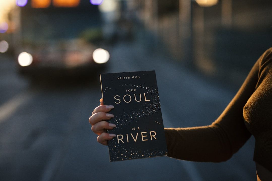 Person, die das Buch „Your Soul is a River“ von Nikita Gill in der Nähe einer unscharfen Stadtstraße in Aachen mit einem Bus im Hintergrund hält.