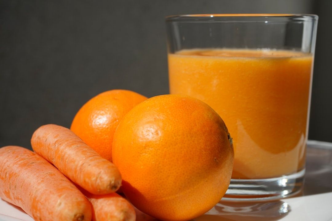 Nahaufnahme eines mit Orangensaft gefüllten Glases, zwei ganzen Orangen und drei Karotten, die neben dem Glas auf einer weißen Oberfläche in einer Aachener Küche platziert sind.