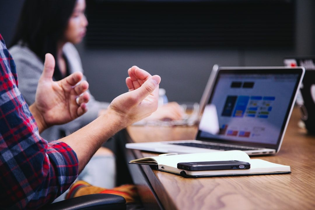 Zwei Personen diskutieren in einem Besprechungsraum. Im Mittelpunkt stehen die gestikulierenden Hände einer Person vor einem geöffneten Laptop, einem Notebook und einem Smartphone, die auf dem Tisch liegen. Im Hintergrund ist ein von Aachen inspiriertes Kunstwerk zu sehen.