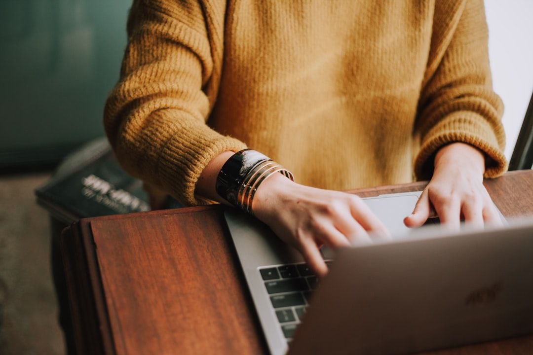 Eine Person in einem senffarbenen Pullover tippt auf einem Laptop, der auf einer Holzoberfläche steht, und arbeitet möglicherweise an ihrer nächsten Reise nach Aachen.