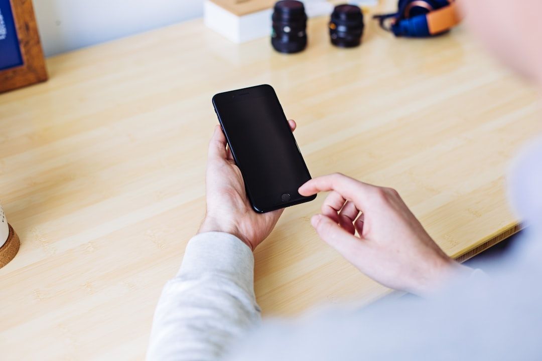 Eine Person in Aachen hält ein Smartphone in der Hand und drückt mit dem Finger auf den Bildschirm, während sie an einem Holzschreibtisch sitzt. Im Hintergrund sind lässig Linsen und Kopfhörer platziert.