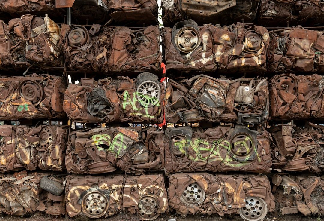 Gestapelte, verrostete und zusammengepresste Autokarosserien bilden eine Wand, die an die Aachener Industrielandschaften erinnert. Räder und verschiedene zerquetschte Teile sind sichtbar, einige davon mit verblassten Graffiti versehen.
