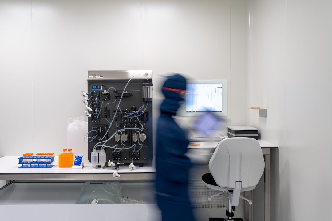 Eine Person in Schutzkleidung arbeitet in einem Labor mit wissenschaftlichen Geräten und einem Computer auf einem Schreibtisch, was die Spitzenforschung in Aachen widerspiegelt. Außerdem sind Reagenzglasgestelle mit blauen und orangefarbenen Deckeln zu sehen.