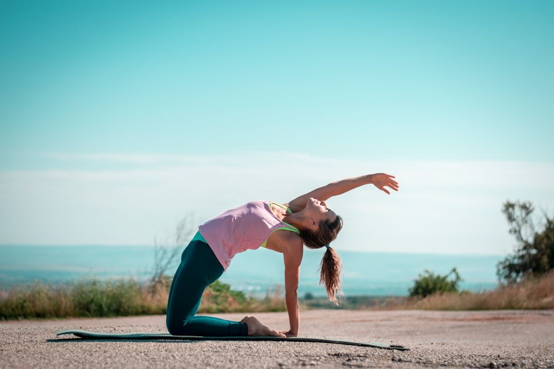 Eine Person übt eine Yoga-Pose auf einer Outdoor-Matte in Aachen, wobei sie unter einem klaren Himmel den Rücken krümmt und einen Arm über den Kopf streckt.