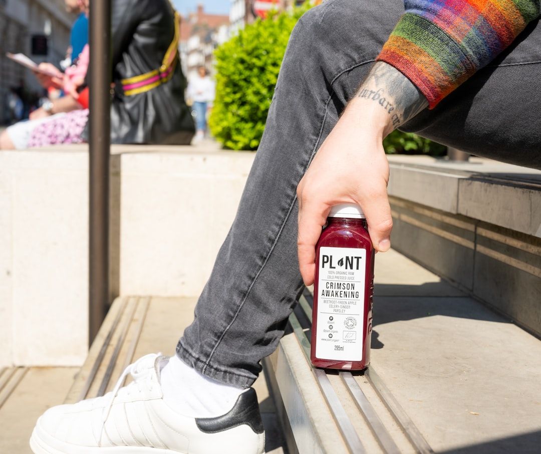 Eine Person in grauen Jeans und buntem Pullover sitzt auf den Stufen des Aachener Doms und hält eine Flasche „Plant“ mit der Aufschrift „Crimson Awakening“ in der Hand.