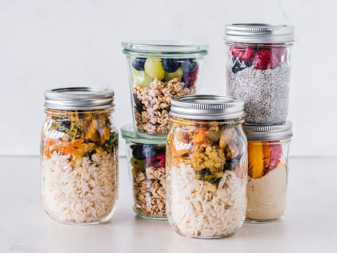 Sechs Gläser, gefüllt mit unterschiedlichen Lebensmitteln, darunter Reis, Müsli, Obst und Chia-Pudding, ordentlich auf einer weißen Fläche angeordnet, erinnern an die Schlichtheit einer Aachener Küche.