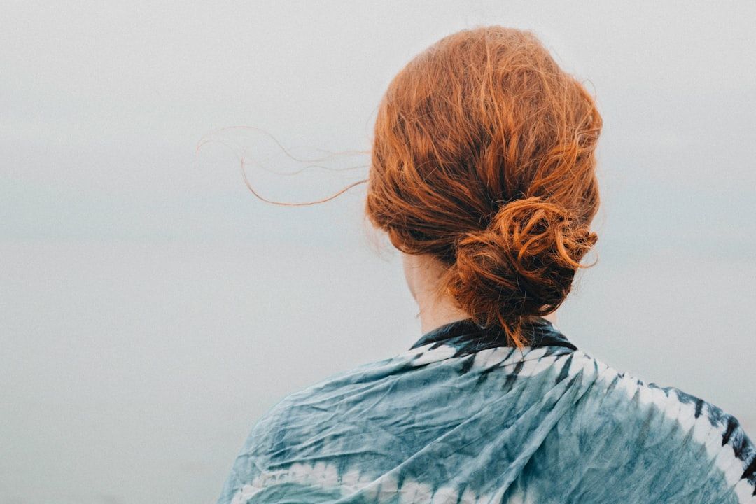 Eine Person mit zurückgebundenem roten Haar und Batik-Shirt steht mit dem Rücken zur Wand und blickt auf eine neblige Landschaft, die an die ruhigen Außenbezirke Aachens erinnert.