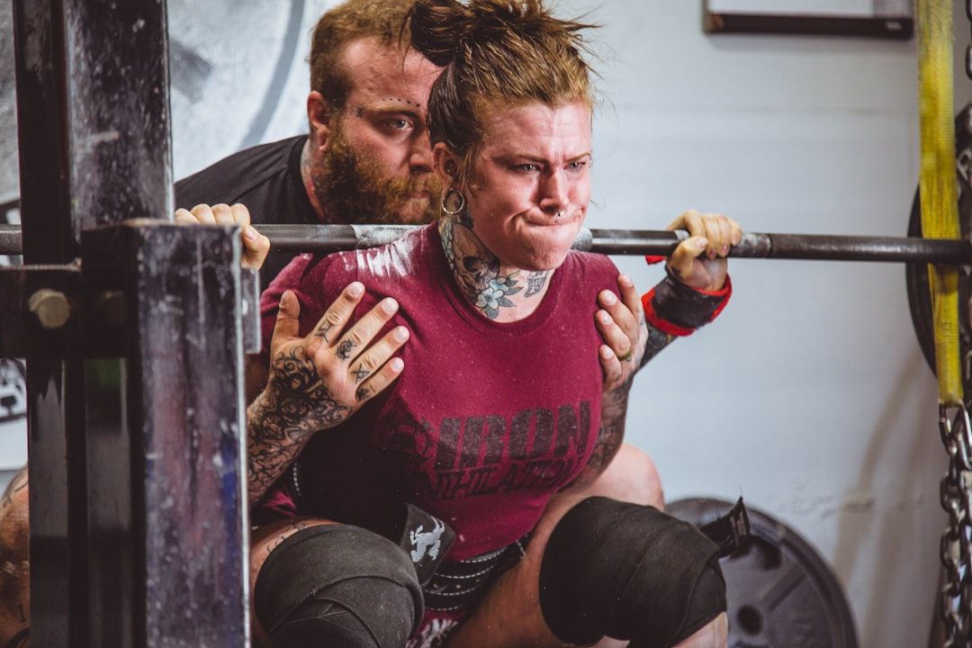 In einem Fitnessstudio in Aachen führt eine tätowierte Person Kniebeugen mit einer Langhantel aus, während eine andere Person sie genau beobachtet.