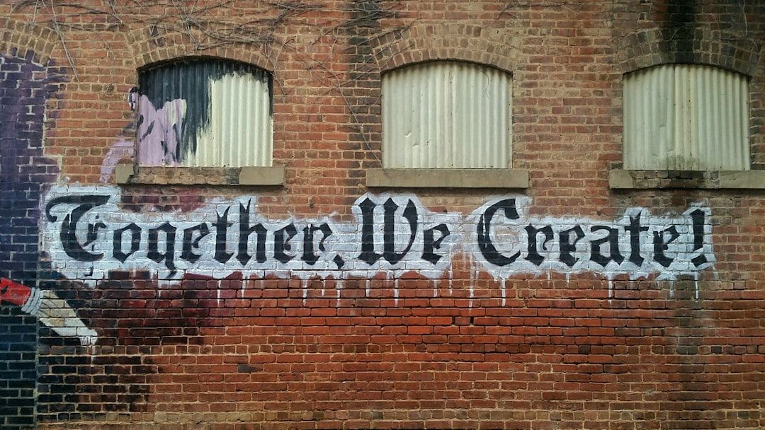 Auf einer Backsteinmauer in Aachen ist der Graffiti-Text „Together, We Create!“ in weißer Farbe mit schwarzer Umrandung zu sehen, links ist ein Teilbild einer Person zu sehen und darüber drei geschlossene Fenster.