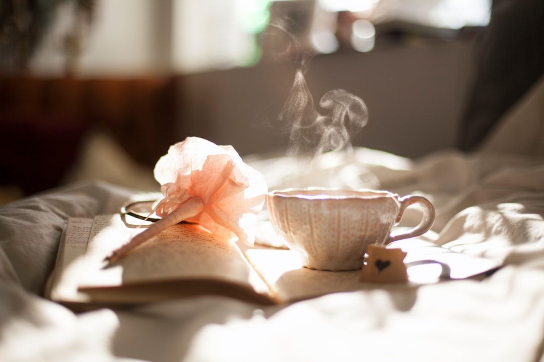 Eine dampfende Tasse Tee, ein offenes Notizbuch und eine rosa Blume stehen auf einem Bett in sanftem Tageslicht und erzeugen eine heitere Atmosphäre, die an einen frühen Morgen in Aachen erinnert.