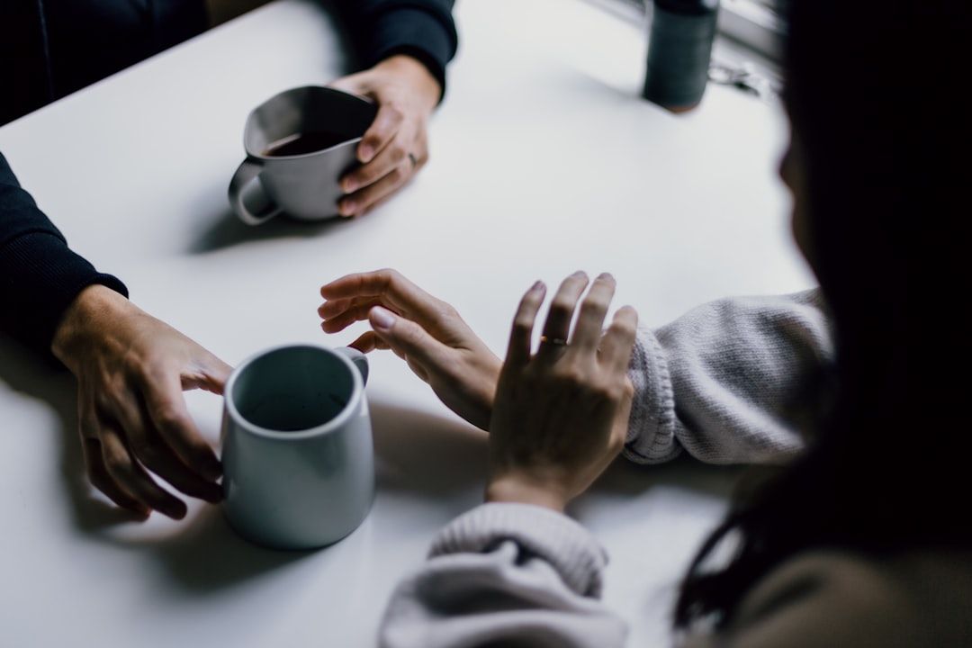 In einem urigen Café in Aachen sitzen sich zwei Menschen an einem Tisch gegenüber, jeder hält einen Becher Kaffee in den Händen. Ihre Hände liegen nah beieinander, was ein vertrauliches Gespräch suggeriert.