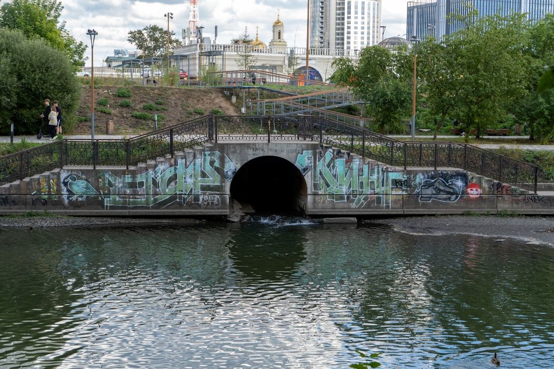 Eine Steinbrücke mit Graffiti überspannt einen Fluss in einem Stadtpark in Aachen, mit Bäumen und historischen Gebäuden im Hintergrund.