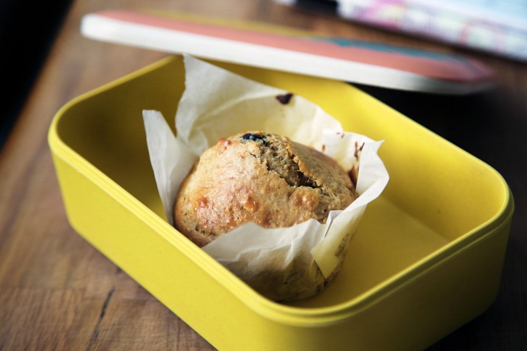 Ein in Backpapier eingewickelter Muffin liegt in einer gelben rechteckigen Form auf einer Holzoberfläche, die an die charmanten Cafés in Aachen erinnert. Der teilweise sichtbare Deckel der Form lehnt sanft dagegen.