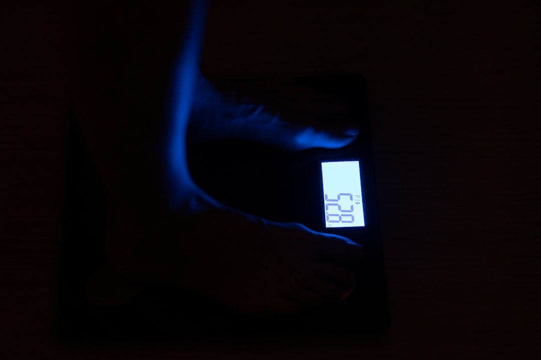 Die Füße einer Person stehen auf einer digitalen Waage, die ein Gewicht von 52,8 anzeigt. Die schwach beleuchtete Szene wirft ein sanftes Licht vom Display und erinnert an einen abendlichen Spaziergang durch Aachens ruhige Straßen.