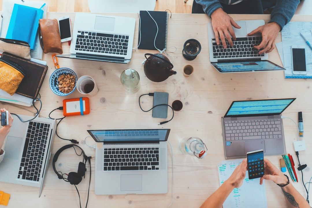 Ein Tisch in Aachen ist mit Laptops, Telefonen, Notizbüchern, Kopfhörern, einer Teekanne und Snacks geschmückt, umgeben von Menschen, die an den Geräten arbeiten.