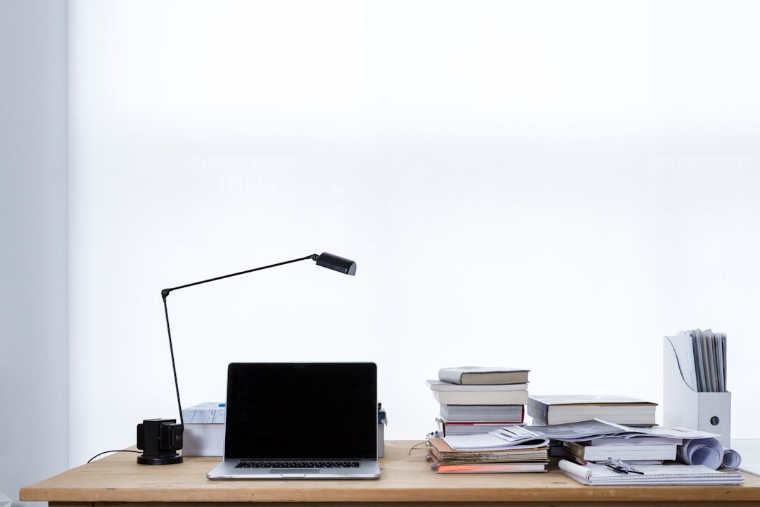 Ein ordentlicher Büroschreibtisch in Aachen mit einem aufgeklappten Laptop, einer Schreibtischlampe, mehreren Stapeln Büchern, Akten und einer Tasse.