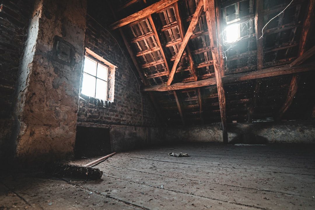 Schwach beleuchtetes Dachgeschoss mit Holzbalken, die an den historischen Charme Aachens erinnern, einem kleinen Fenster, das Licht hereinlässt, unverputzten Ziegelwänden und einem staubigen Holzboden.