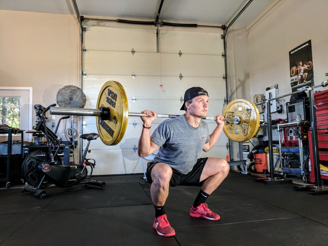 Eine Person in grauem T-Shirt und schwarzen Shorts macht in einem Aachener Fitnessstudio eine Kniebeuge mit einer Hantelstange. Im Hintergrund sind Trainingsgeräte und ein großes Garagentor zu sehen.