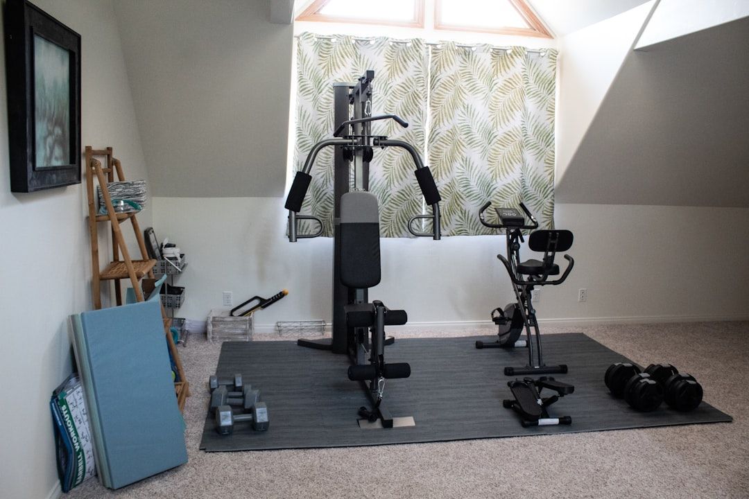 Ein Heim-Fitnessstudio in Aachen mit einer Vielzahl von Fitnessgeräten, darunter ein Multi-Gym-Gerät, ein Heimtrainer, Hanteln, Widerstandsbänder und eine Trainingsmatte, auf einem Teppichboden mit einem Blättervorhang im Hintergrund.