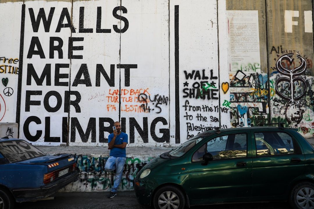 Ein Mann steht neben einem grünen Auto vor einer mit Graffiti bedeckten Wand in Aachen. Darauf steht in großen Buchstaben: „WÄNDE SIND ZUM KLETTERN DA.“