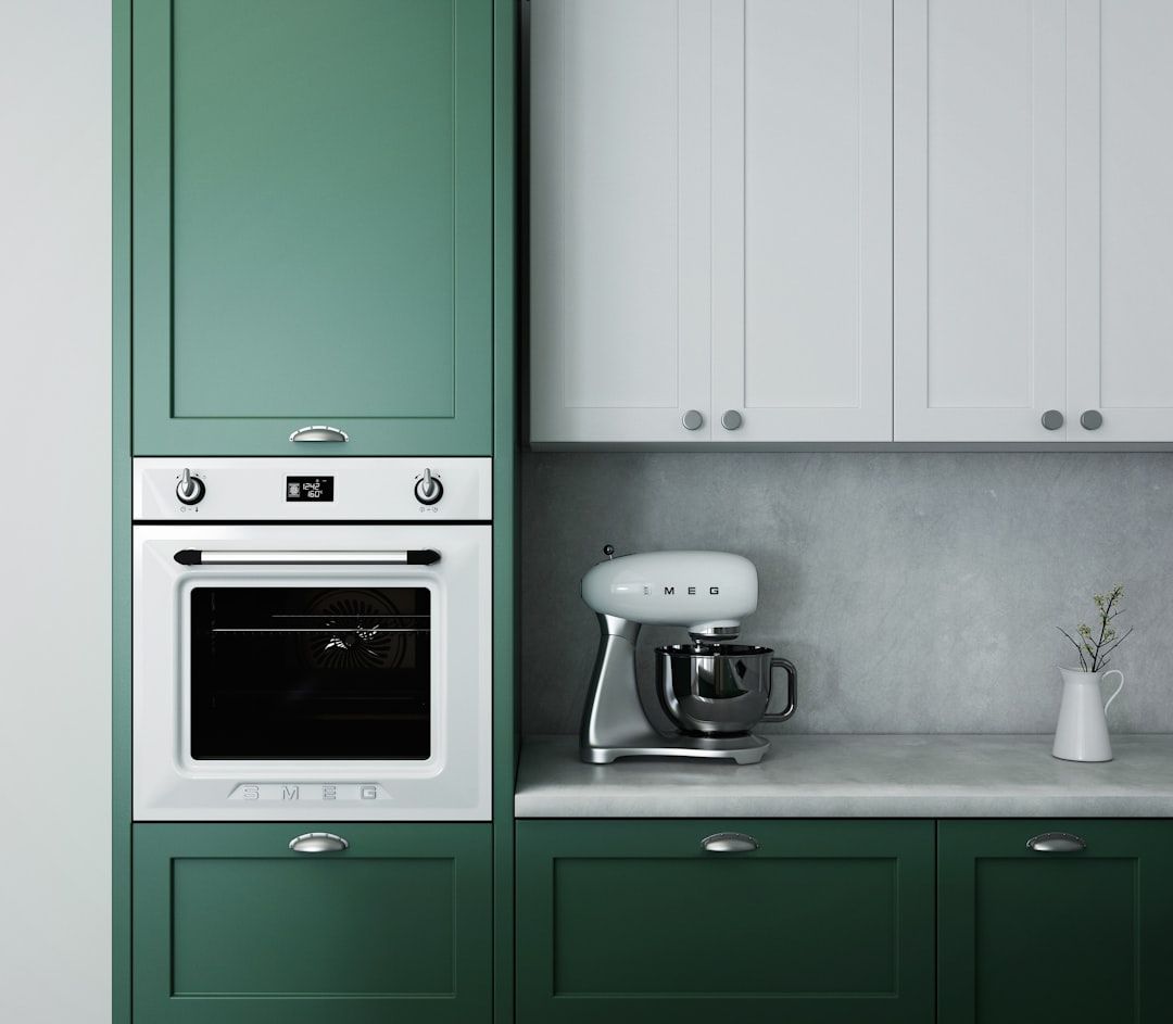 Eine moderne Küche mit grünen Unterschränken, weißen Oberschränken, die an das schlichte Design Aachens erinnern, einem Einbaubackofen, einer Standmixer-Maschine und einer kleinen weißen Vase mit einem Zweig.