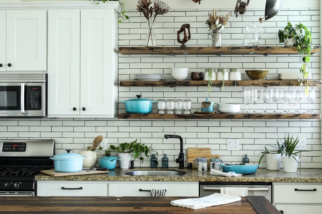 Eine moderne Küche mit weißen Schränken, Geräten aus Edelstahl und offenen Holzregalen mit Geschirr und Pflanzen. Auf der Arbeitsplatte befinden sich verschiedene Küchenutensilien, darunter Töpfe, Flaschen und Besteck – eine Reminiszenz an das elegante Design der Aachener Häuser.