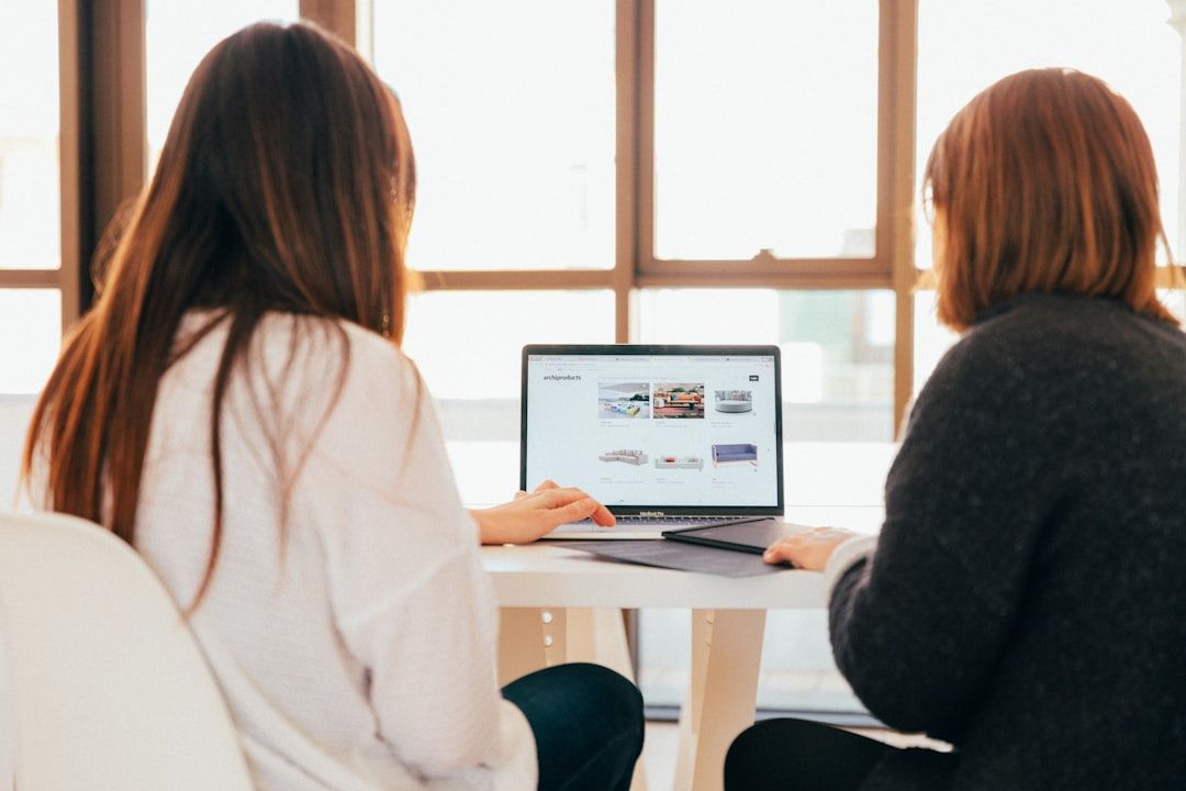 Zwei Frauen sitzen an einem Tisch in Aachen und schauen auf den Bildschirm eines Laptops, auf dem verschiedene Miniaturbilder von Webseiten angezeigt werden. Im Hintergrund sind große Fenster zu sehen, durch die Sonnenlicht fällt.