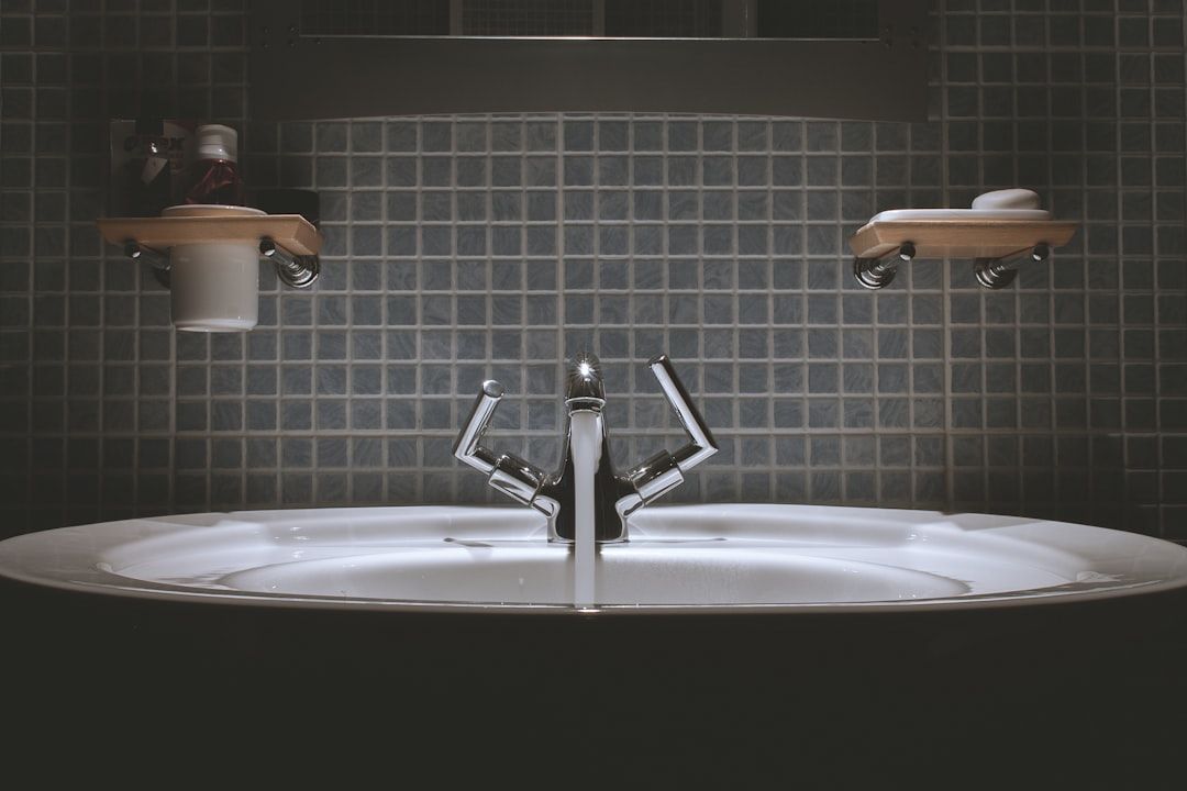 Ein modernes Waschbecken mit einem Zweigriff-Wasserhahn, umgeben von einer gefliesten Rückwand, die an den klassischen Charme Aachens erinnert, und Regalen über dem Waschbecken, auf denen verschiedene Toilettenartikel aufbewahrt werden.