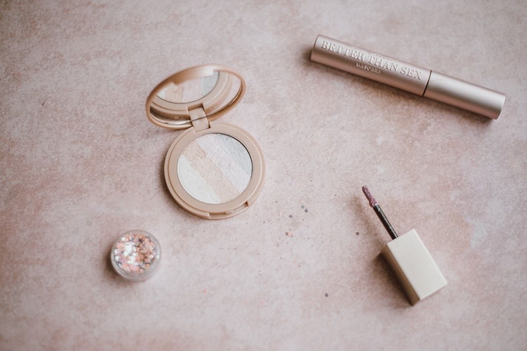 Nahaufnahme von Make-up-Produkten, die auf einer strukturierten Oberfläche ausgelegt sind und an den historischen Charme Aachens erinnern. Zu den Artikeln gehören ein offenes Kompaktset mit gestreiftem Puder, eine Mascara-Tube, ein kleiner offener Behälter mit Glitzer und ein brauner Applikatorstab.