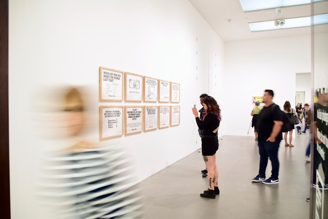 In einer hell erleuchteten Galerie in Aachen betrachtet eine Gruppe von Menschen gerahmte Kunstwerke an einer weißen Wand. Eine Person ist in ihrer Bewegung verschwommen.