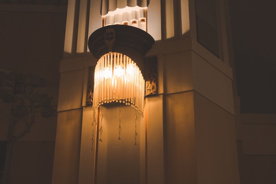Eine verzierte Wandleuchte mit Fransendesign strahlt ein warmes Licht aus, beleuchtet einen Teil der Wand und erinnert an die detaillierten architektonischen Merkmale, die an den historischen Charme Aachens erinnern.