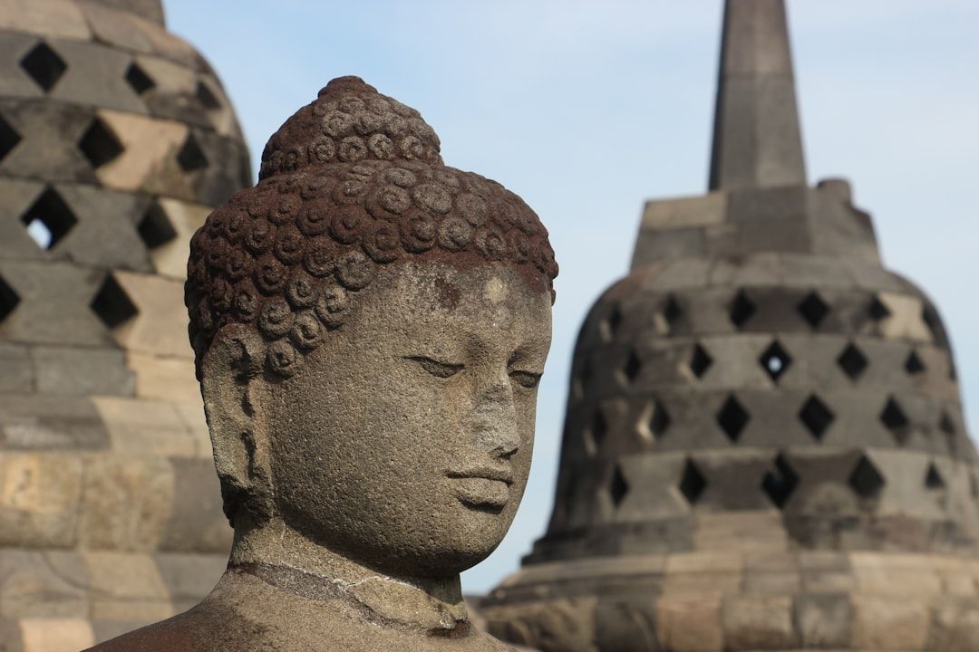 Nahaufnahme einer ruhigen Buddha-Statue aus Stein mit zwei alten Stupa-Strukturen im Hintergrund vor einem klaren Himmel, die an das historische Ambiente Aachens erinnert.