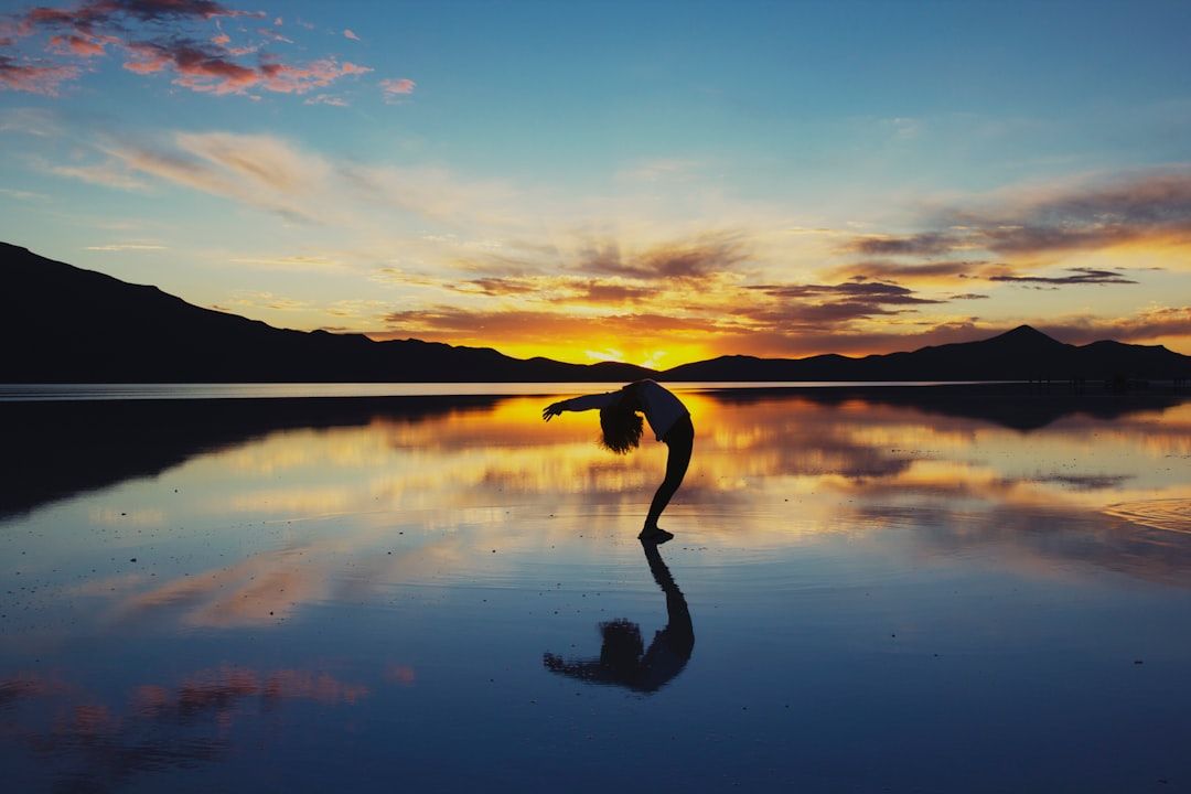 Eine Person führt bei Sonnenuntergang einen Rückwärtsbogen auf einer reflektierenden Oberfläche aus, mit den majestätischen Aachener Bergen und einem bunten Himmel im Hintergrund.
