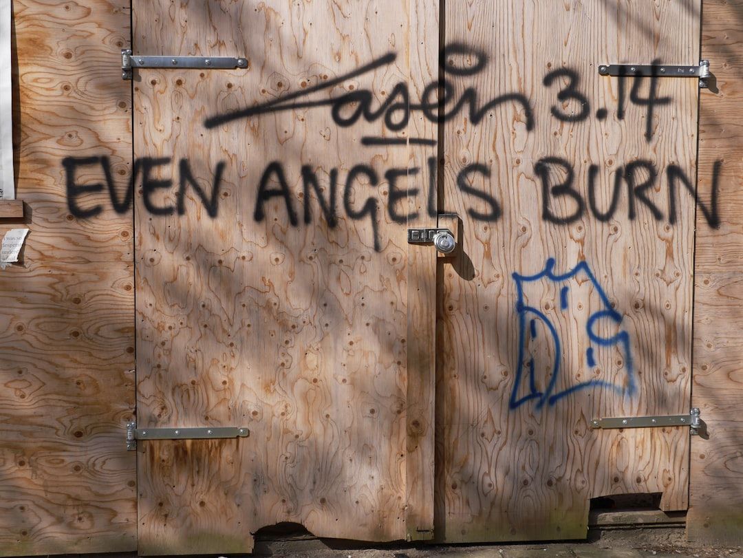 Eine Holztür mit Metallscharnieren in Aachen ist mit dem Text „EVEN ANGELS BURN“ und weiteren Graffiti in schwarzer Farbe besprüht.