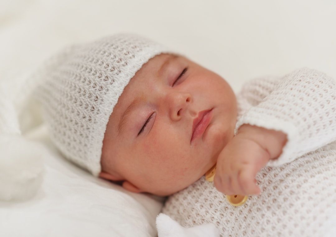 Ein schlafendes Baby in einem weißen Strickoutfit und einer passenden Mütze liegt auf einer weißen Decke und erinnert an die heitere Atmosphäre der ruhigen Morgenstunden Aachens.