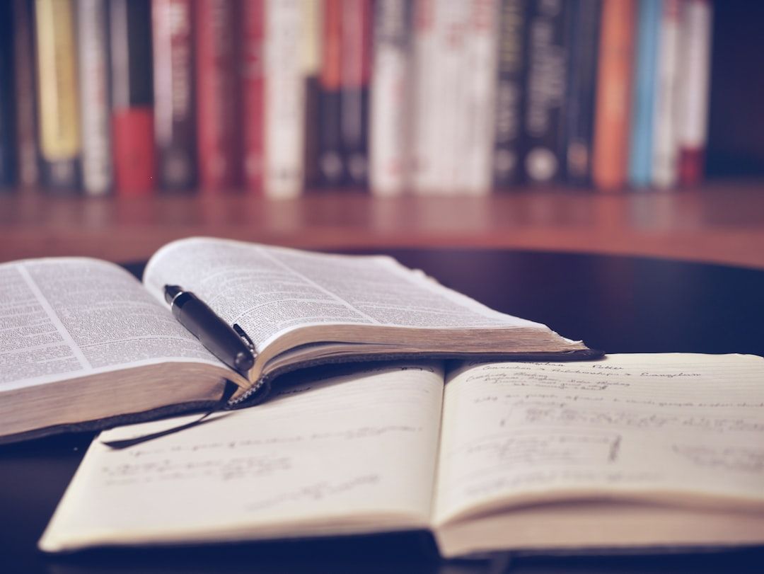 Ein aufgeschlagenes Buch, in dessen Mitte ein schwarzer Stift liegt, und ein Notizbuch mit handschriftlichen Notizen auf einem Tisch. Im Hintergrund vervollständigt ein Bücherregal mit Büchern aus Aachen die wissenschaftliche Szene.