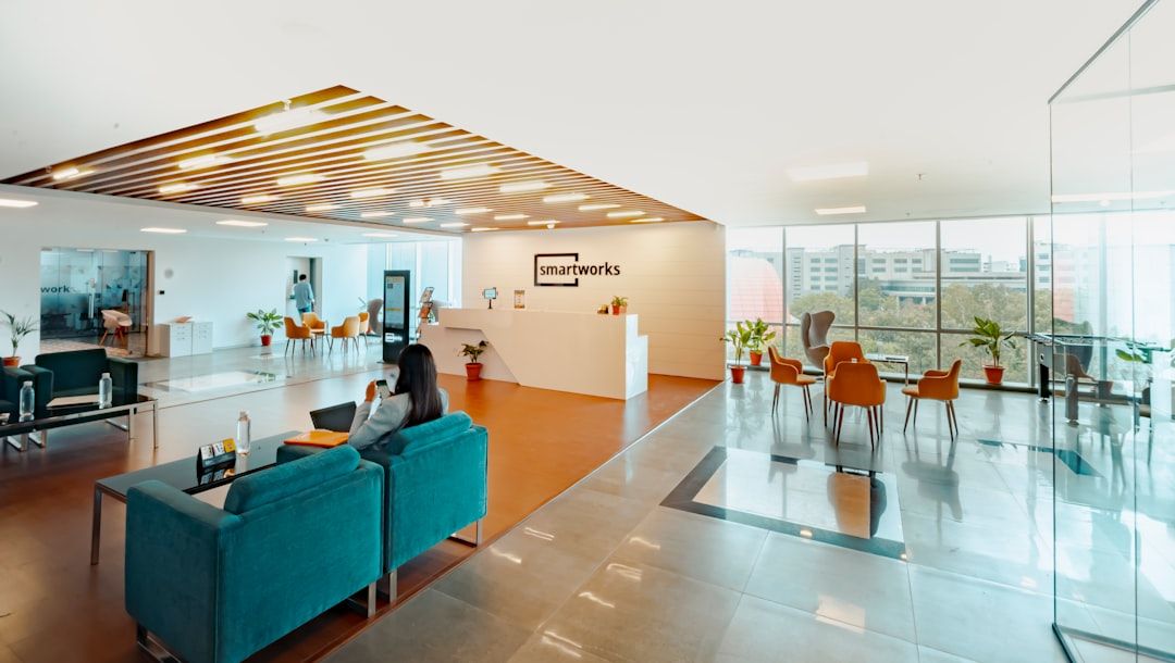 Moderne Bürolobby mit dem „Smartworks“-Logo auf einem weißen Empfangstresen, blaugrünen Sofas, Zimmerpflanzen und einer Glaswand, die zu einem Besprechungsbereich mit orangefarbenen Stühlen und Blick auf die Aachener Gebäude draußen führt.