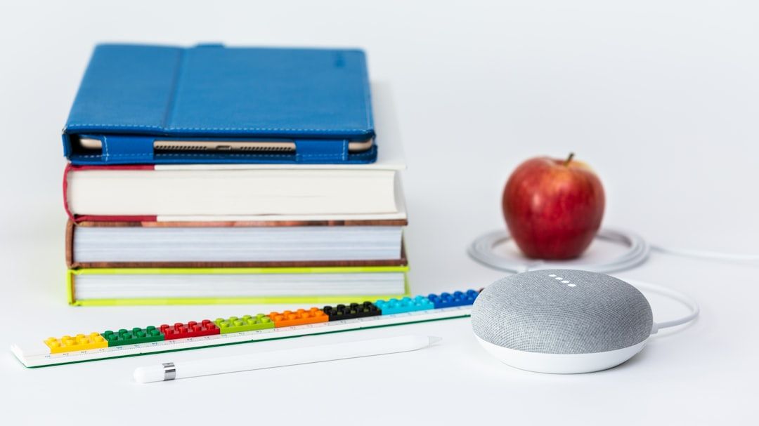 Ein Stapel Bücher mit einem blauen Einband obenauf, ein Apfel, ein weißer Stift, ein buntes Lineal und ein grauer Smart Speaker sind auf einer weißen Oberfläche angeordnet und erinnern an eine akademische Einrichtung in Aachen.
