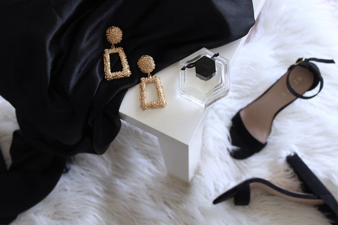 Goldene Ohrringe, ein geöffnetes Parfümfläschchen, schwarze High Heels und ein schwarzer Stoff werden auf einer weißen Oberfläche und einem flauschigen weißen Teppich präsentiert und erinnern an die schicke Raffinesse der Aachener Modeszene.