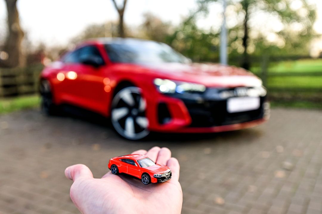 Hand hält ein rotes Spielzeugauto vor einem verschwommenen roten Auto, das auf einer Aachener Einfahrt mit Bäumen im Hintergrund geparkt ist.