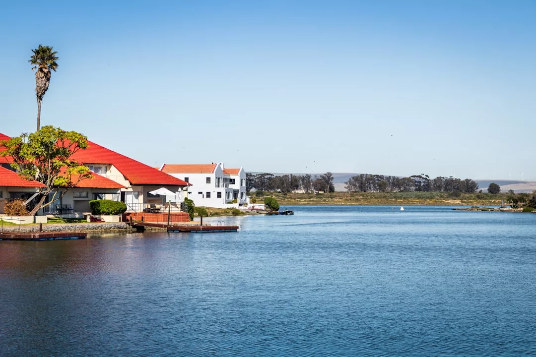 Seeuferszene mit Häusern, darunter eines mit rotem Dach, einer Palme und ruhigem Wasser unter einem klaren blauen Himmel.