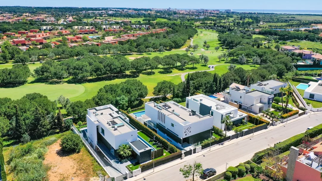 Luftaufnahme moderner weißer Villen mit Flachdächern und grünen Rasenflächen in der Nähe eines Golfplatzes und eines Waldgebiets mit einem fernen Horizont.